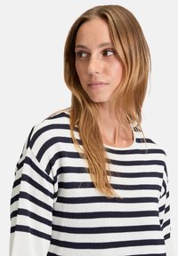 Frau mit hellbraunem Haar, die ein weißes und dunkelblaues, horizontal gestreiftes Langarmshirt trägt und nach links schaut, vor einem schlichten Hintergrund.