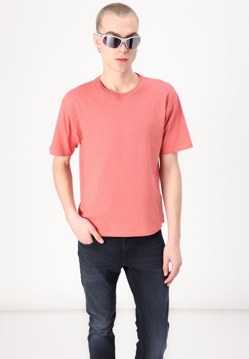 T-shirt en coton corail à manches courtes et col rond, associée à un jean en denim foncé. Le mannequin porte des lunettes de soleil gris clair avec des verres foncés.