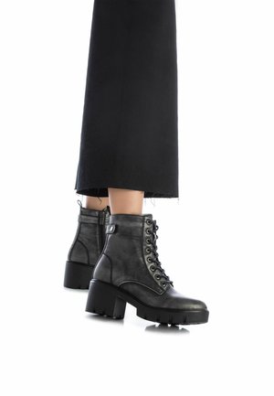 Botas de cuero negro con cordones hasta el tobillo y tacones gruesos, llevadas con pantalones negros cortos sobre un fondo blanco.