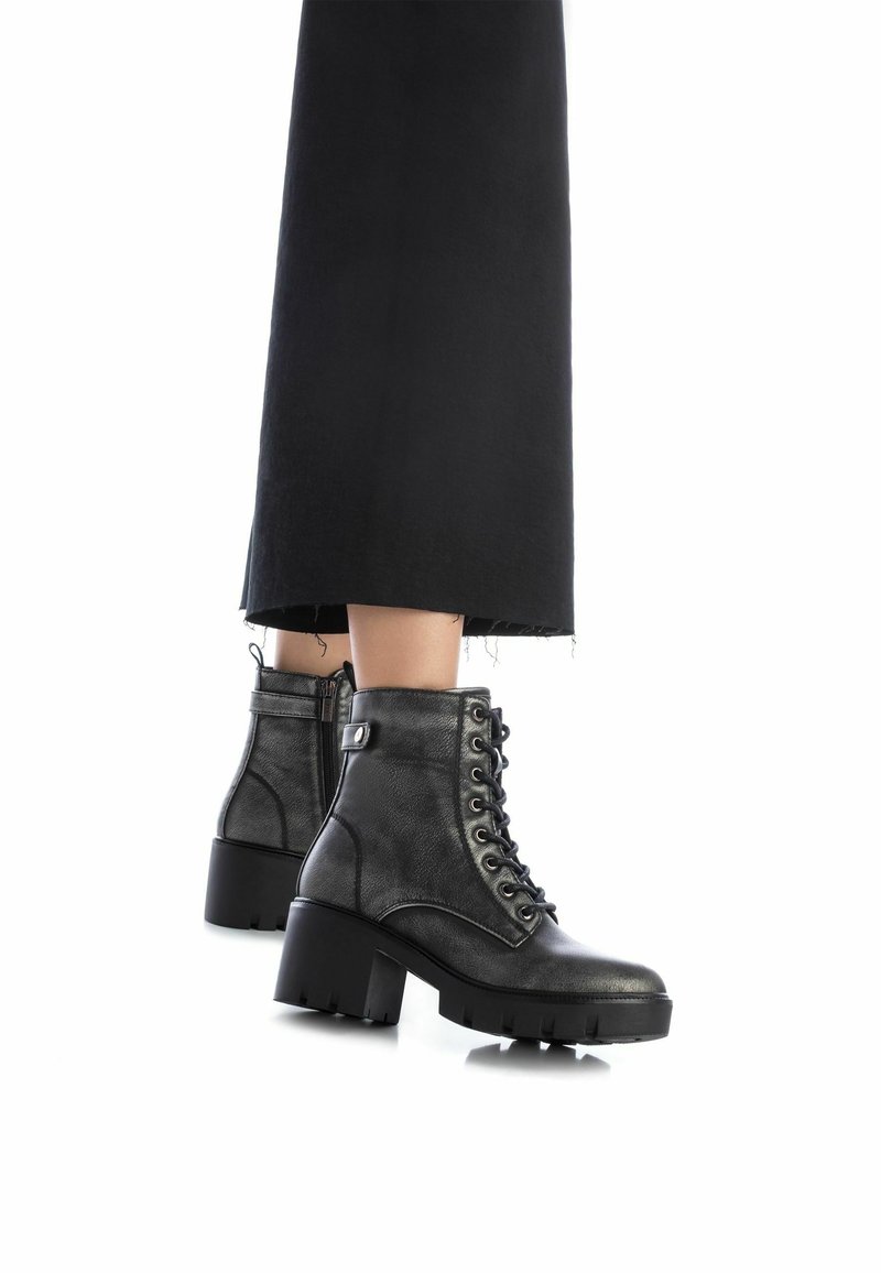 Botas de cuero negro con cordones hasta el tobillo y tacones gruesos, llevadas con pantalones negros cortos sobre un fondo blanco.