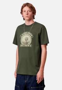 Camiseta de algodón verde con un estampado gráfico que presenta el texto "Carhartt WIP Detroit". Las mangas cortas y un corte holgado completan el diseño.
