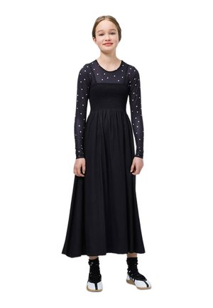 Zwarte jurk met lange, nauwsluitende mouwen met polka dots, een gerimpeld lijfje en een vloeiende rok. Draagt zwarte en witte sneakers.