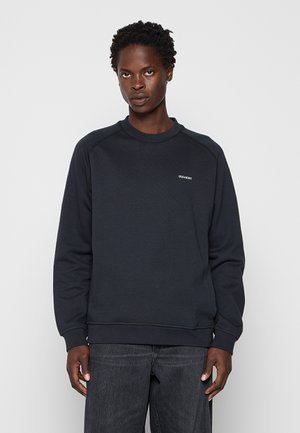 Mann trägt schwarzen Sweatshirt mit Rundhalsausschnitt und kleinem weißen „LYLE & SCOTT“-Logo auf der Brust sowie dunkelgraue Jeans vor weißem Hintergrund.