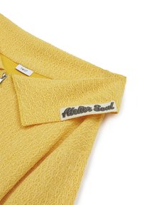 Veste zippée texturée jaune avec un large col, arborant une étiquette "TWIST" et un patch "Atelier Soul" sur le col.