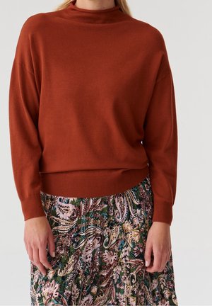 Kvinde iført en langærmet sweater i rustfarve og et plisseret skørt med grønne, rosa og beige blomstrede paisley-mønstre.