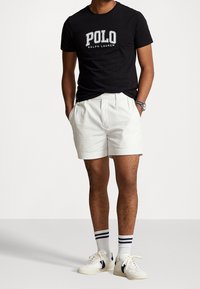 Muž v černém tričku Polo Ralph Lauren, bílých skládacích šortkách, bílých ponožkách s černými pruhy a bílých teniskách s černými akcenty.