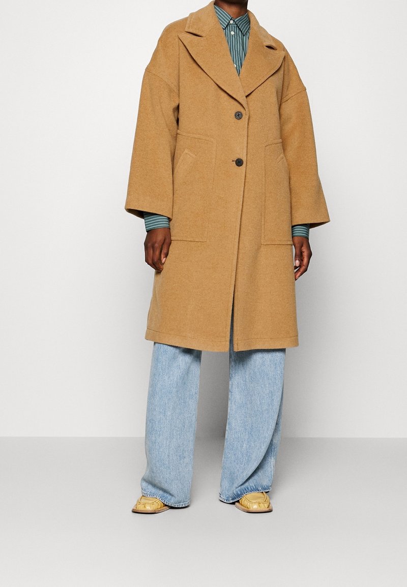 Personne portant un manteau en laine couleur camel avec deux boutons, une chemise rayée, un jean bleu clair à jambes larges et des mocassins beiges, debout devant un fond uni.