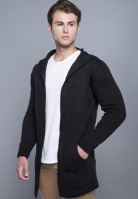 Sort strikket cardigan med hætte, lange ærmer, åben front og to forlommer. Båret over en hvid t-shirt og beige bukser.