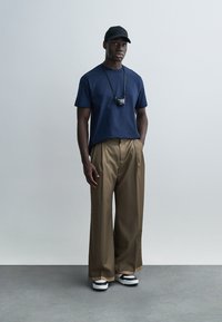 Μπλούζα σε ναυτικό μπλε, μπεζ φαρδιά παντελόνια, μαύρο καπέλο, μαύρο κολιέ με μενταγιόν και μαύρα και λευκά παπούτσια. Μινιμαλιστικός σχεδιασμός με άνετη εφαρμογή.