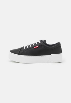 Zapatillas sintéticas negras con una suela de goma blanca, con cordones blancos y un pequeño logo rojo de Levi's en el costado. Diseño de corte bajo.