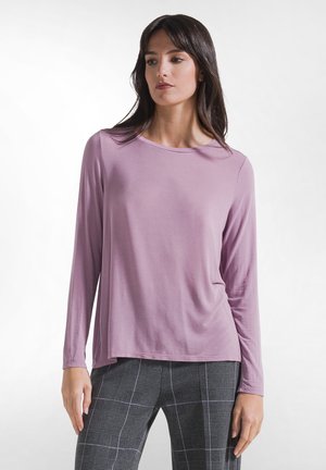 Vrouw die een lavendelkleurige top met lange mouwen en grijze geruite broek draagt, staand tegen een effen witte achtergrond.