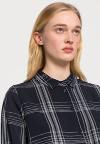 Camisa azul marinho com padrão quadriculado branco, de design abotoado e colarinho, com uma textura de tecido suave e mangas compridas.