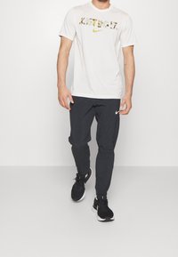 Biała koszulka Nike z logo w wzór kamuflażu, zestawiona z czarnymi zwężanymi dresami i czarnymi butami sportowymi, stojąca na jednolitym tle.