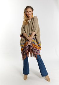 Poncho bej cu modele geometrice colorate și ciucuri; asortat cu blugi evazați și pantofi cu toc bej.