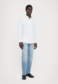 Hvit langermet skjorte med krage og knapper, kombinert med lyseblå straight-leg jeans. Modellen har på seg svarte sko. Enkelt design.