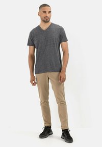 Grijze v-hals t-shirt gemaakt van textuurstof; gecombineerd met lichtbruine broeken en zwarte schoenen. Model staat in een ontspannen houding tegen een witte achtergrond.