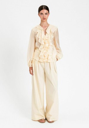 Donna in piedi che indossa una camicetta crema con volant e maniche lunghe e pantaloni palazzo, abbinati a sandali bassi, su sfondo bianco.