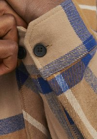 Beige geruite flanellen overhemdmanchet met blauwe en witte accenten, voorzien van twee zwarte knopen en een zachte, gestructureerde stof.