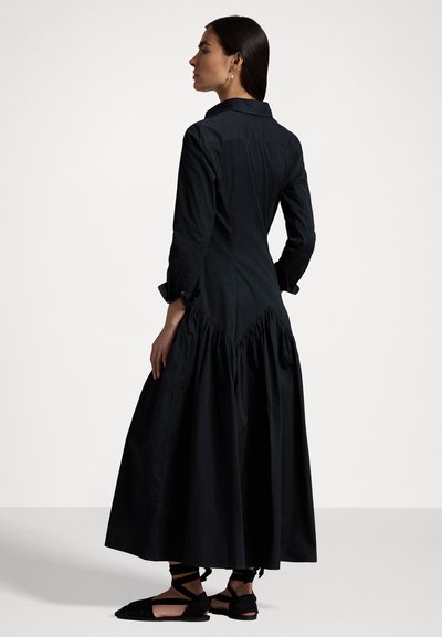Zwarte katoen jurk met een aansluitende bodice, lange mouwen, een kraag en een volumineuze, gathered rok. Draagt met zwarte sandaaltjes met bandjes.