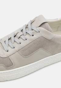 Zapatilla gris clara que combina cuero suave y gamuza. Suela de goma texturizada y cordones planos grises. Marca sutil en el costado.