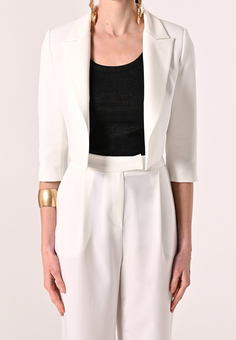 Witte getailleerde blazer over een zwarte top met structuur, gecombineerd met witte broeken met hoge taille. Met een gouden armband en grote oorbellen.