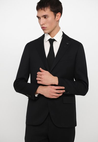 Traje negro a medida con solapa muesca, bolsillo único, diseño ajustado y textura de tela sutil. Combinado con una camisa blanca y una corbata negra.