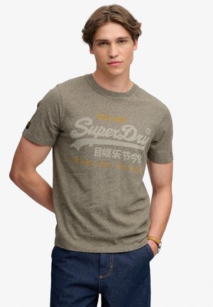 Jeune homme portant un t-shirt marron de la marque Superdry et un jean bleu, debout avec une main dans la poche devant un fond uni.