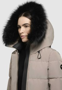 Világos bézs színű puffasztott kabát fekete műszőrme kapucnival. Elöl zipzárral, állítható húzózsinórral és logóval ellátott folttal a ruhaujján.