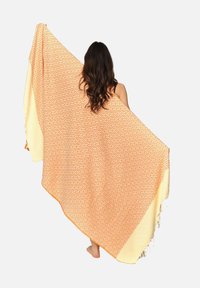 Stor orange og cremefarvet mønstret tæppe med diamantformer og flosset kant, draperet over en person. Lavet af blødt tekstilmateriale.