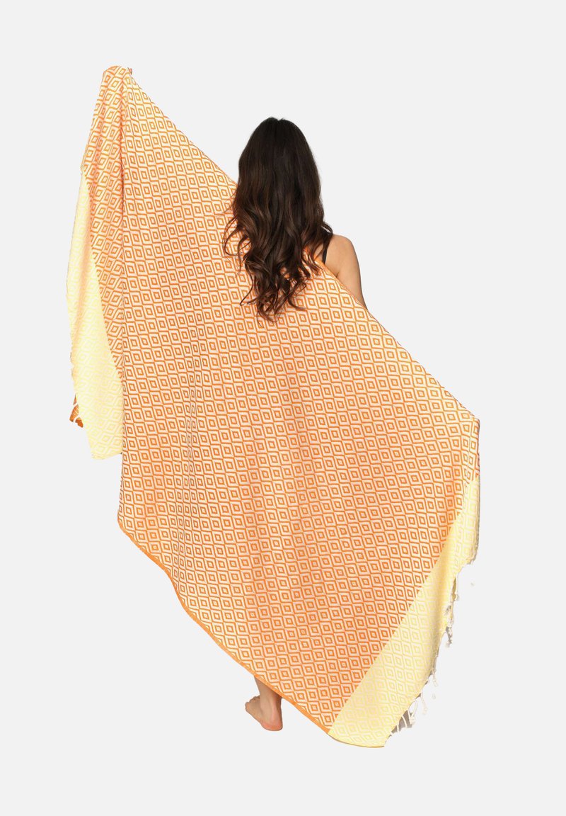 Stor orange og cremefarvet mønstret tæppe med diamantformer og flosset kant, draperet over en person. Lavet af blødt tekstilmateriale.