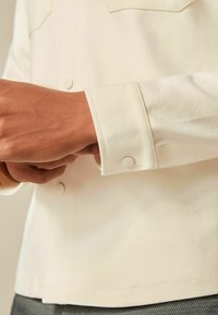 Nærbillede af en person, der justerer cuffen på en cremedejakke med knapdetaljer over grå bukser.