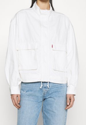 Personne portant une veste blanche à fermeture éclair avec de grandes poches avant et un jean bleu clair, debout contre un fond blanc uni.