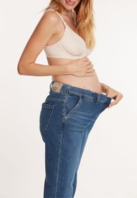Jeans de maternité en denim foncé avec une coupe décontractée, dotés d'une large ceinture pour plus de confort. Un soutien-gorge d'allaitement beige clair est porté en dessous.