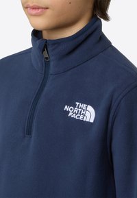 Mørk marineblå fleecegenser med glidelås ved halsen. Har en hvit brodert logo på venstre bryst. Myk tekstur og standard passform.