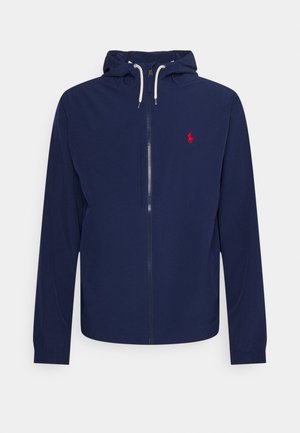 Marineblauwe capuchonjas met volledige ritssluiting aan de voorkant, witte trekkoorden en een klein rood geborduurd logo op de linkerbovenborst.