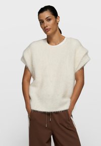 Mujer de cabello oscuro recogido en una coleta baja, lleva un suéter sin mangas blanco y esponjoso sobre una camisa blanca y pantalones marrones, con las manos en los bolsillos.