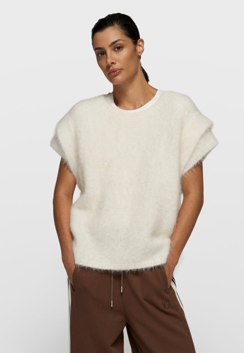 Mujer de cabello oscuro recogido en una coleta baja, lleva un suéter sin mangas blanco y esponjoso sobre una camisa blanca y pantalones marrones, con las manos en los bolsillos.