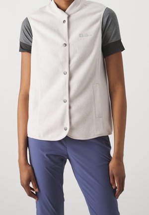 Person trägt weiße Fleeceweste mit Druckknöpfen über grauem kurzärmligem Shirt und blauer Hose, steht mit locker an den Seiten hängenden Armen.