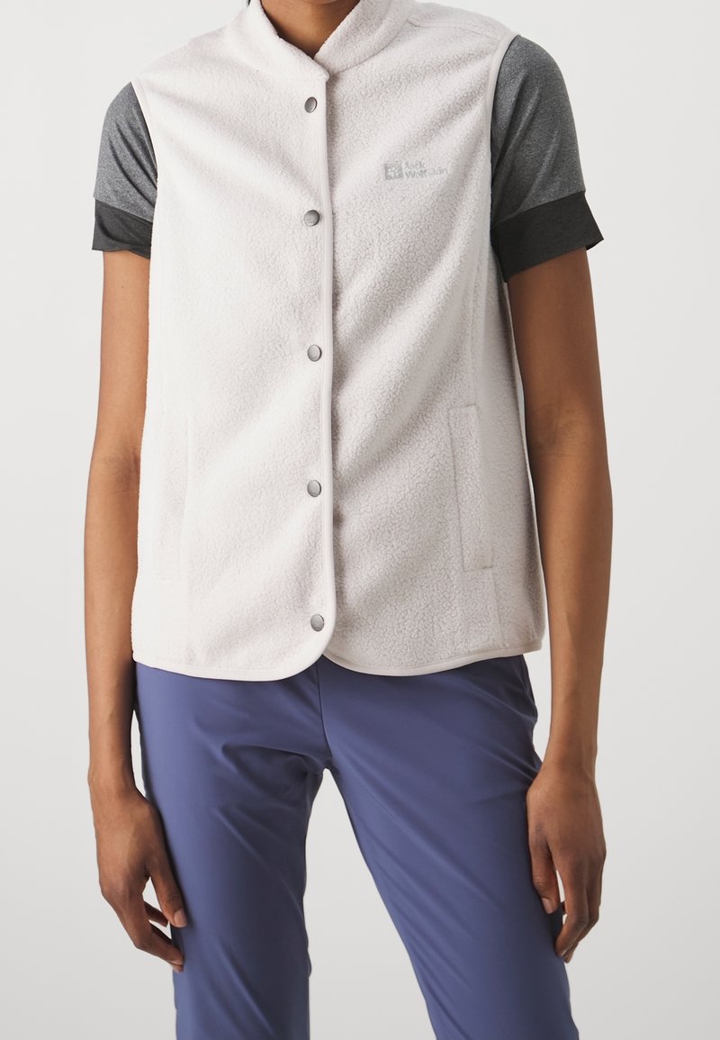 Persona que lleva un chaleco polar blanco con botones de presión sobre una camiseta gris de manga corta y pantalones azules, de pie con los brazos relajados a los lados.
