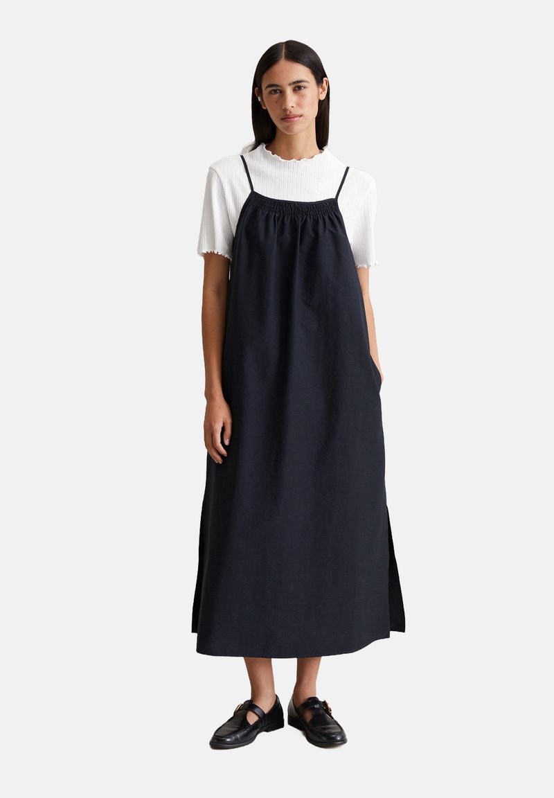 Marineblaues Leinenkleid mit verstellbaren Spaghetti-Trägern, gerafftem Ausschnitt, Seitentaschen und lockerer Passform, über einem weißen gerippten Oberteil getragen.