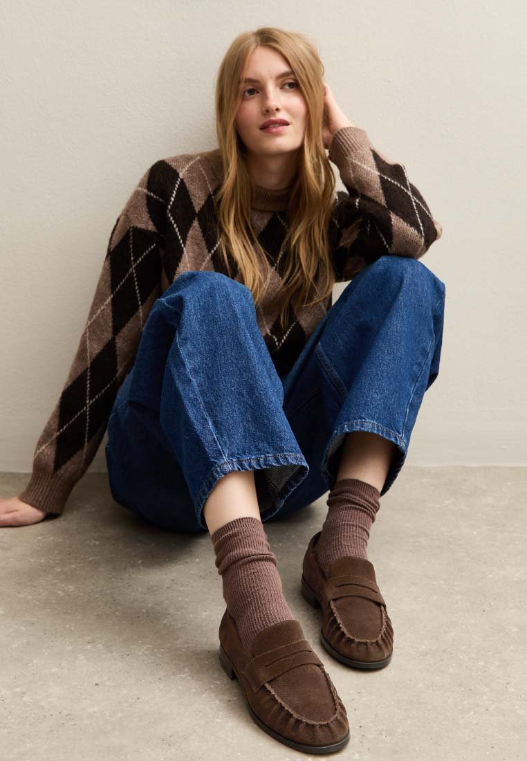 Maglione a motivo argyle marrone, jeans a gamba larga blu, mocassini marroni e calze a coste abbinate. Posizione seduta su uno sfondo neutro.