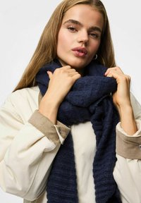 Écharpe en tricot bleu marine avec un design texturé et côtelé, enroulée autour du cou, associée à une veste de couleur claire avec un col en velours côtelé.