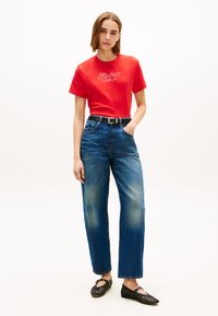 Κόκκινο βαμβακερό μπλουζάκι με το λογότυπο «TOMMY HILFIGER CO.», ψηλόμεσο μπλε τζιν παντελόνι, μαύρη ζώνη και μαύρα πλεκτά πάνινα παπούτσια. Λευκό φόντο.