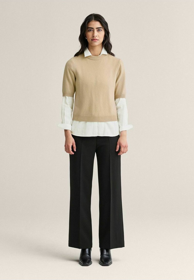 Beige neulottu villapaita lyhyillä hihoilla, joka on kerroksittain valkoisen kauluspuseron päällä, yhdistettynä mustiin löysiin housuihin ja mustiin nilkkureihin.