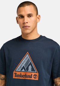 Námořnická modrá bavlněná tričko s trojúhelníkovou grafikou v oranžové a šedé, s nápisem "Timberland" a logem stromu. Krátké rukávy, kulatý výstřih.