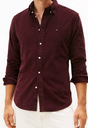 Mand iført en bordeaux knappet skjorte med lange ærmer og hvide knapper, ærmerne er rullede op, kombineret med lyse bukser.