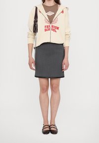 Donna che indossa una felpa crema con zip e il testo rosso "Fashion School", un top marrone, una gonna grigia fino al ginocchio e scarpe Mary Jane scure, con una piccola borsa a tracolla.