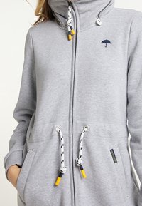 Žena v světle šedé mikině na zip s kapucí, která má žluté šňůrky a malou námořnickou deštníkovou logo na hrudi.