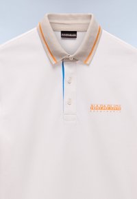 Valkoinen polo-paita, jossa on teksturoitu kangas, beige kaulus, oranssit ja siniset korostusraidat sekä oranssi brodeerattu logo rinnassa.