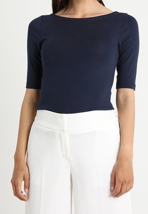 Femme portant un haut bleu marine ajusté à manches jusqu'au coude et un pantalon blanc taille haute à jambes larges, debout avec les bras détendus.